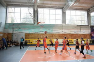 Юные баскетболисты в оранжевой и бордовой форме играют у кольца в спортивном зале