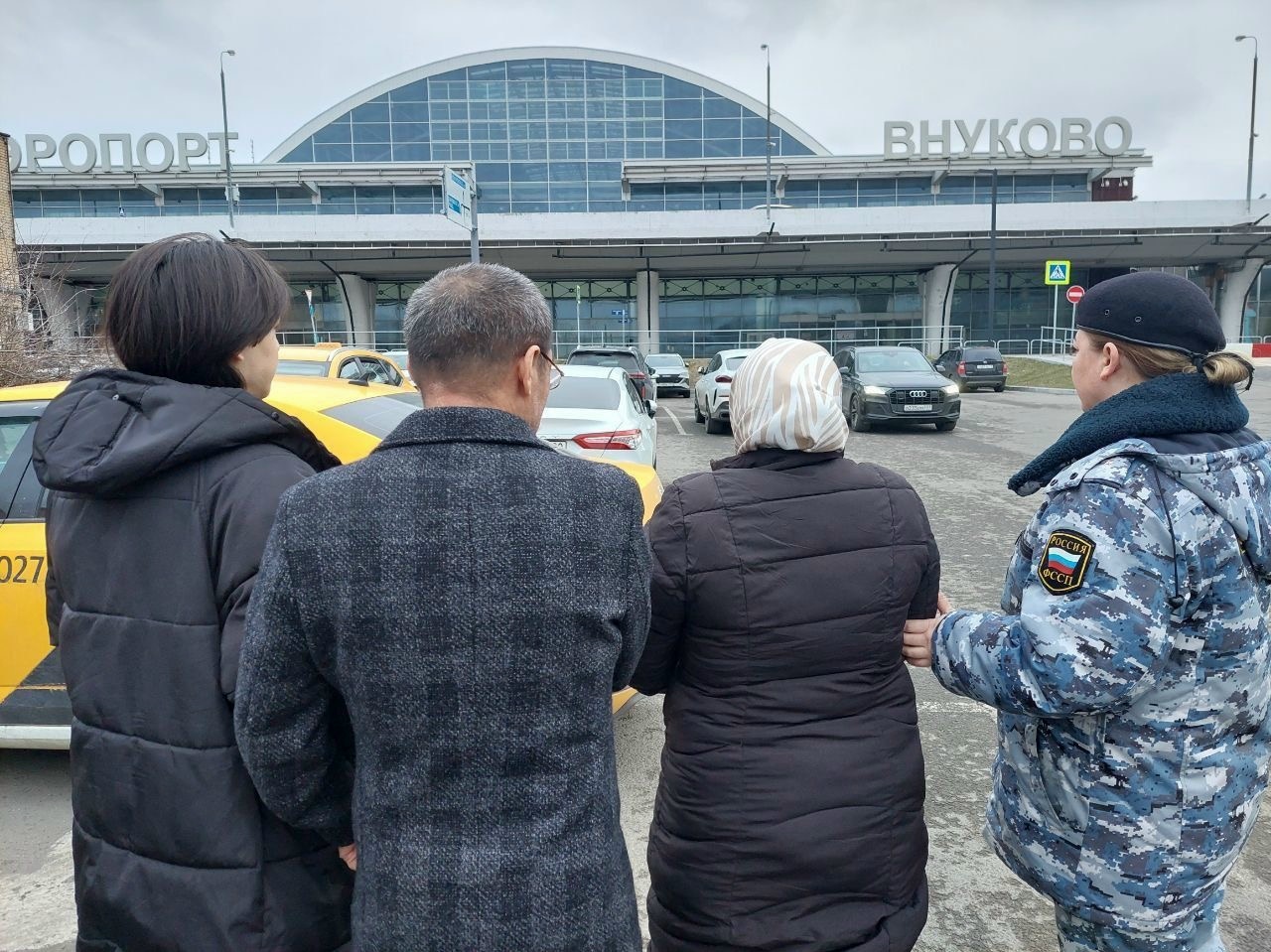 Сотрудница ФССП ведёт задержанную женщину и ещё несколько человек на парковке на фоне здания аэропорта с надписью «ВНУКОВО»
