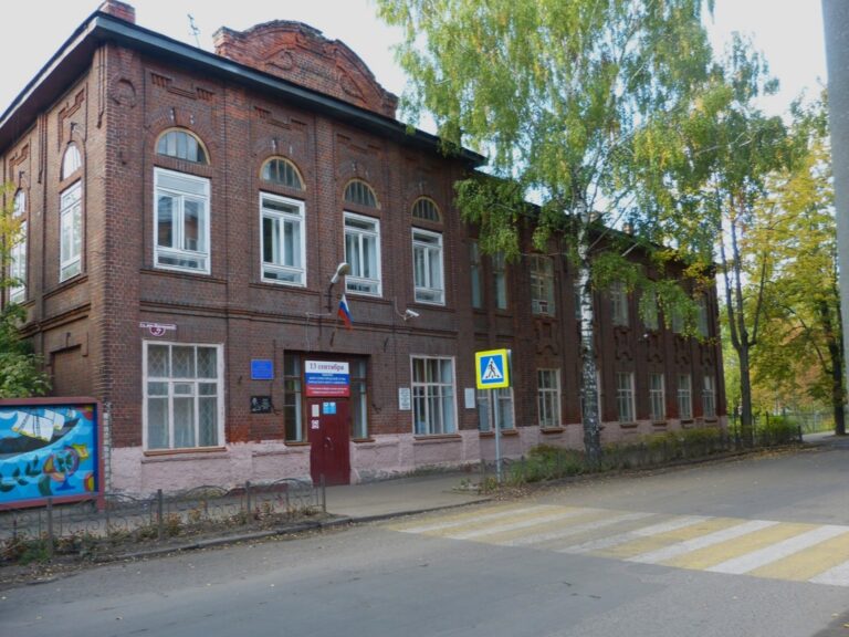 Двухэтажное кирпичное здание гимназии у дороги с пешеходным переходом и берёзами