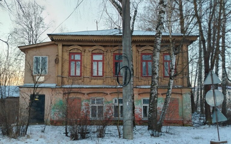 Двухэтажное старое деревянное здание с красными рамами окон, зима, берёзы и столб на переднем плане.