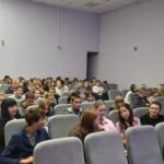 Зрители в небольшом зрительном зале: ряды серых кресел, в них сидят люди разных возрастов в актовом/культурно‑досуговом центре.