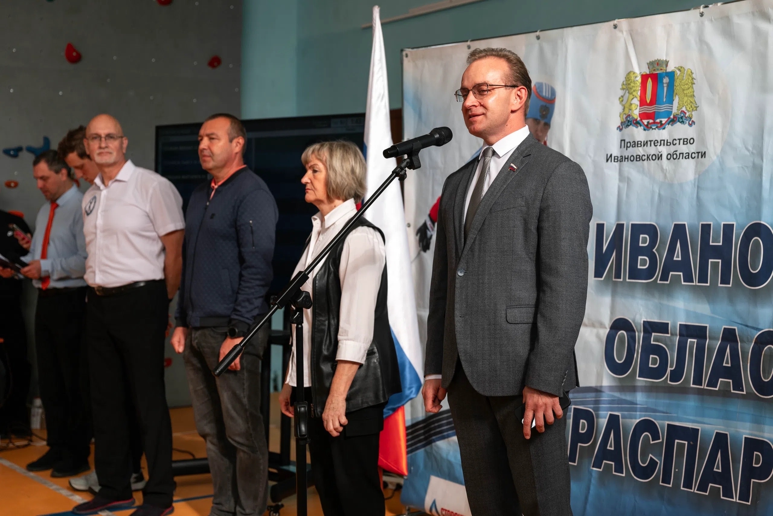 Церемония открытия: несколько взрослых стоят у микрофона на фоне флага и баннера с гербом и надписью «Правительство Ивановской области» (внутренний спортивный зал, скалодром на стене)
