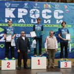 Призёры забега на пьедестале перед большим баннером «Кросс нации»: спортсмены с медалями и дипломами на открытой сцене в лесопарковой зоне.