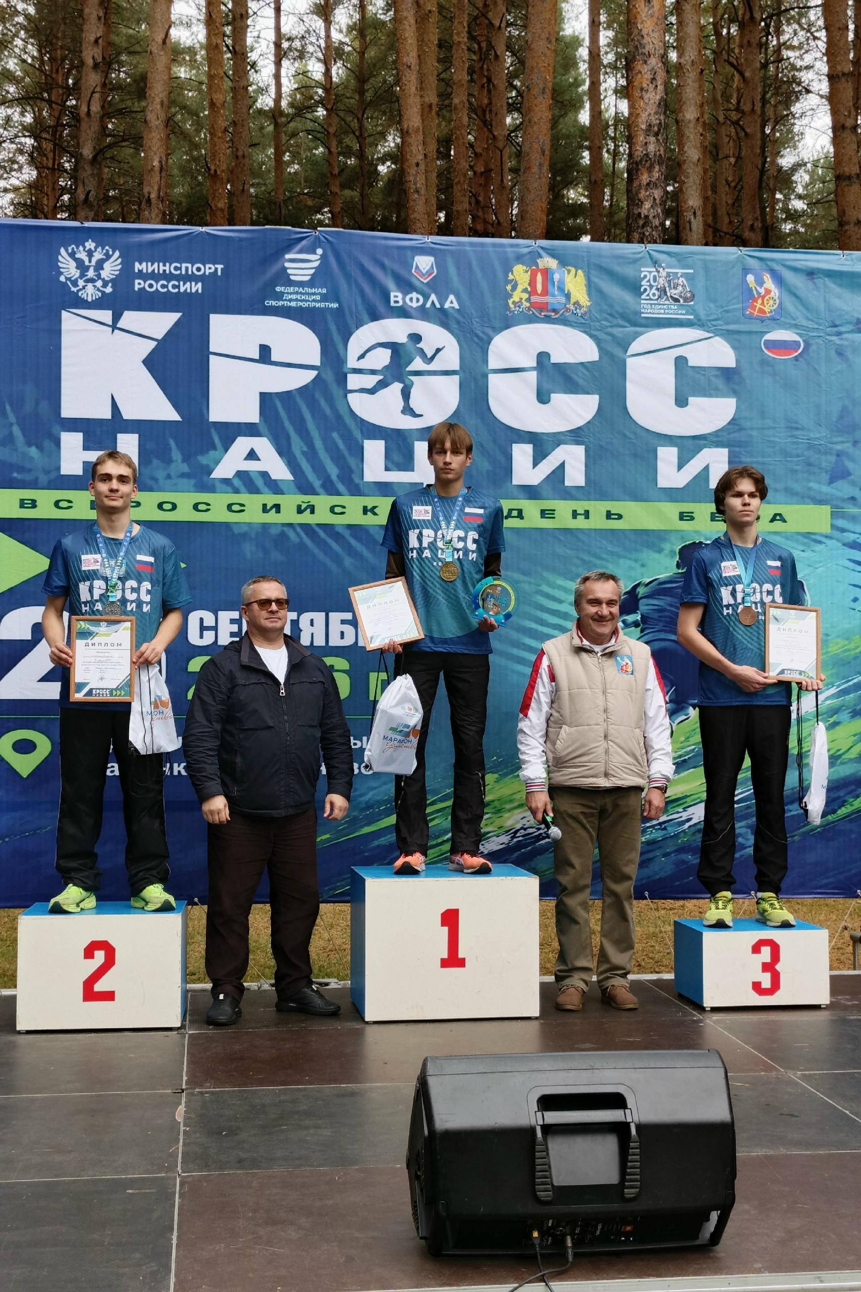 Призёры забега на пьедестале перед большим баннером «Кросс нации»: спортсмены с медалями и дипломами на открытой сцене в лесопарковой зоне.