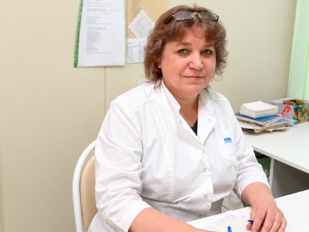 Женщина в белом медицинском халате сидит за столом с документами в кабинете