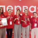 Пять девушек в одинаковых красных футболках на фоне красного баннера с надписью «первая», держат медали, диплом и стеклянный приз