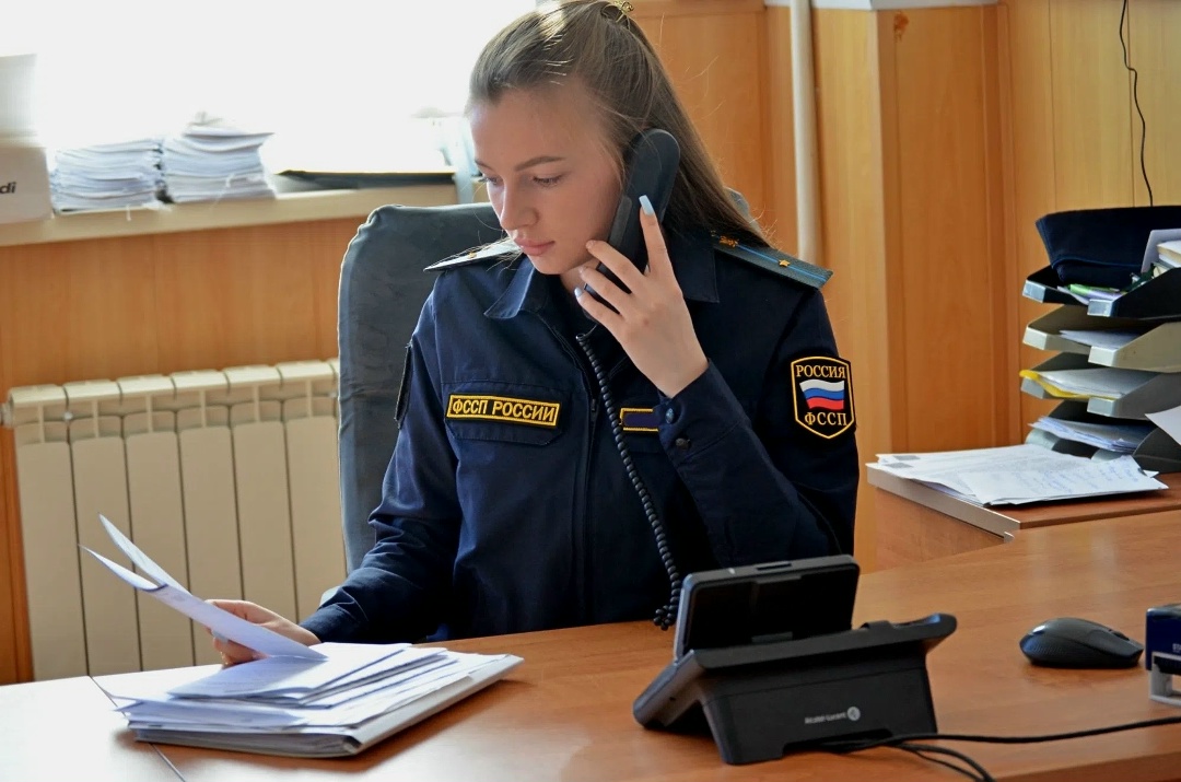 Сотрудница в форме ФССП России за рабочим столом держит телефон и просматривает бумаги.