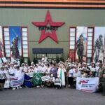 Групповая фотография участников и организаторов военно‑спортивного мероприятия у здания с большой красной звездой и надписью «ПАТРИОТ», люди в белых футболках и кепках, несколько флагов и плакатов с логотипом «Защитники Отечества».
