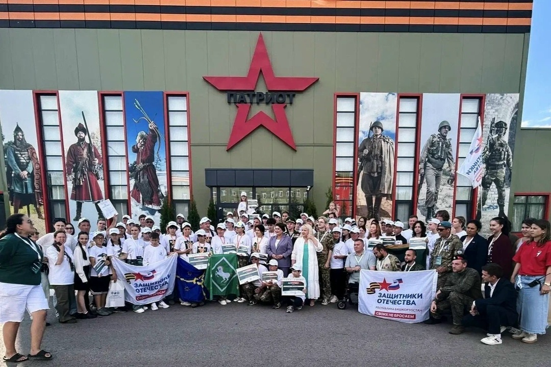Групповая фотография участников и организаторов военно‑спортивного мероприятия у здания с большой красной звездой и надписью «ПАТРИОТ», люди в белых футболках и кепках, несколько флагов и плакатов с логотипом «Защитники Отечества».