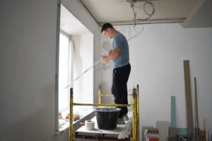 Мужчина на строительных лесах держит длинный металлический профиль в комнате с незаконченной проводкой