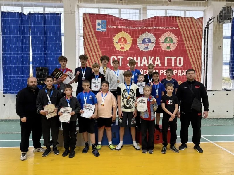 Группа юных спортсменов и два тренера в спортивном зале: медали на шее, дипломы в руках, победный подиум с цифрами 1–3 на фоне баннера.