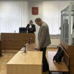 Судебный зал: судья за трибуной, человек у стола и подсудимые в металлической клетке справа