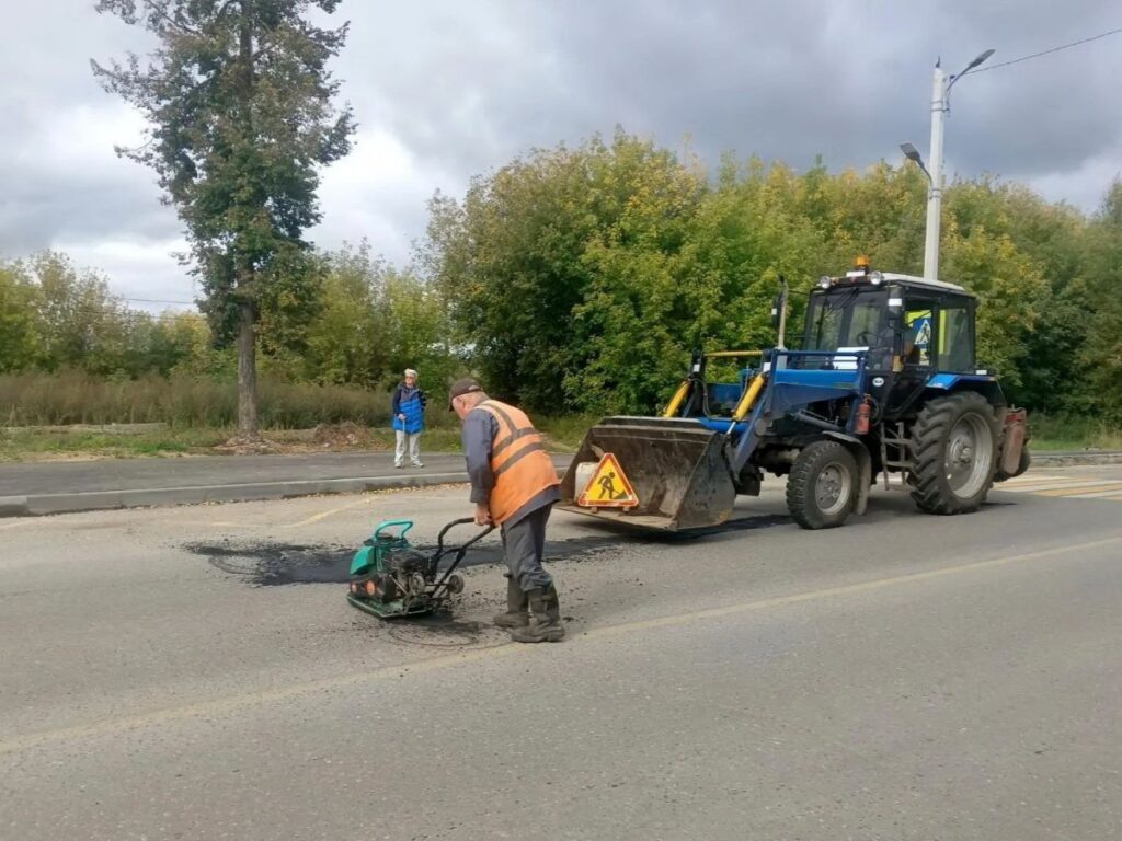 Рабочий в оранжевом жилете уплотняет асфальт виброплитой рядом с синим трактором у пешеходного перехода