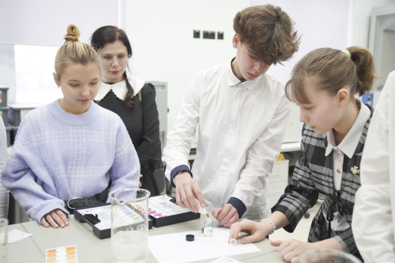 Подростки и преподаватель в учебной лаборатории за столом с колбами, пипетками и реактивами.