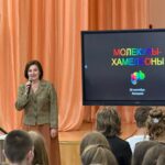 Лектор с микрофоном на сцене перед большим экраном, на экране заголовок «Молекулы-хамелеоны», в зале видны школьники.