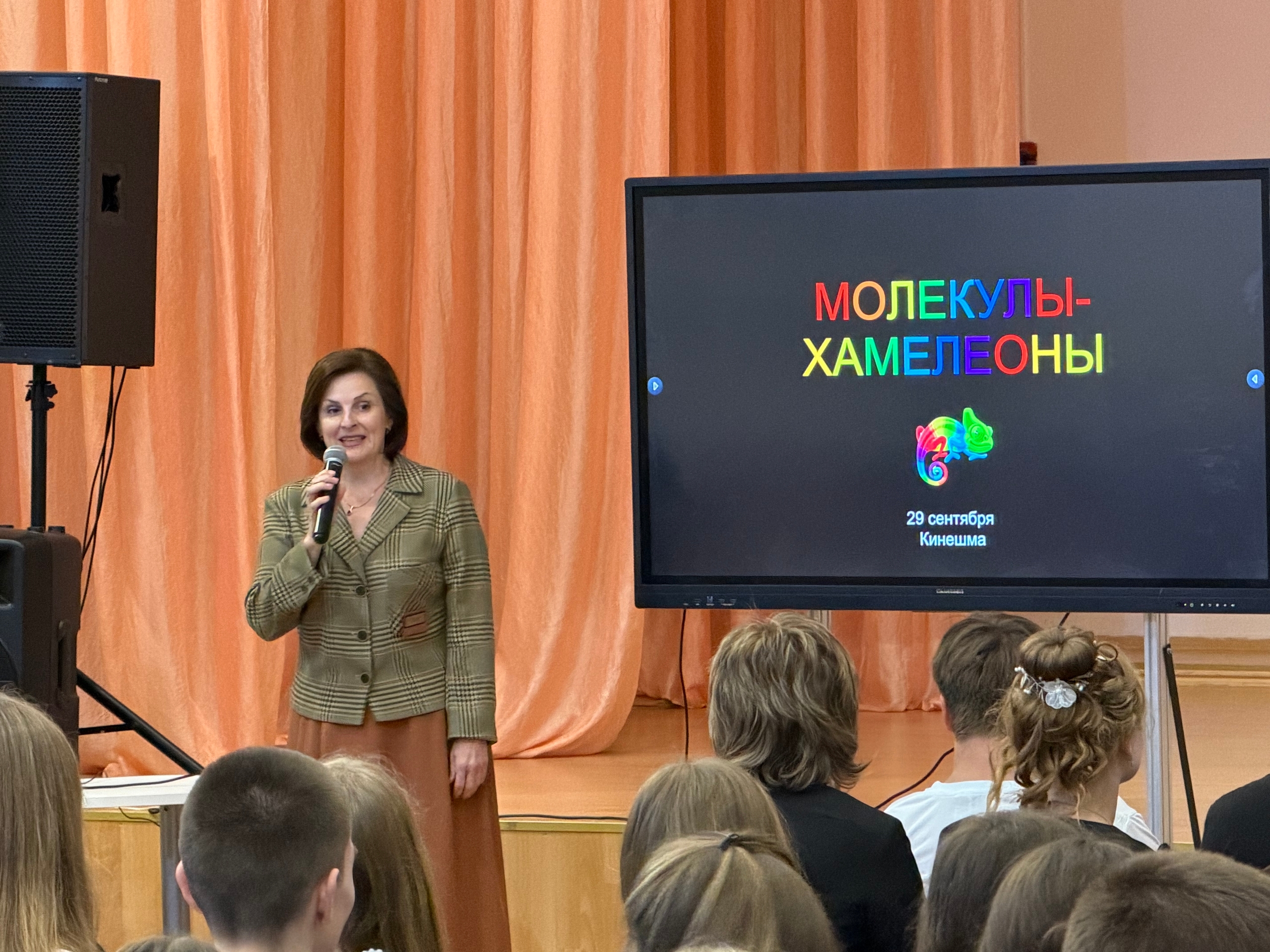 Лектор с микрофоном на сцене перед большим экраном, на экране заголовок «Молекулы-хамелеоны», в зале видны школьники.