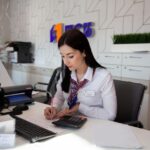 Сотрудница банка за рабочим столом в офисе, заполняет документы и считает на калькуляторе; на заднем плане виден логотип банка.