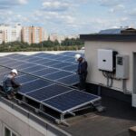 На крыше многоквартирного дома ряд солнечных панелей; два монтажника в защитных касках работают рядом; на стене видны инвертор и открытый электрический щит.