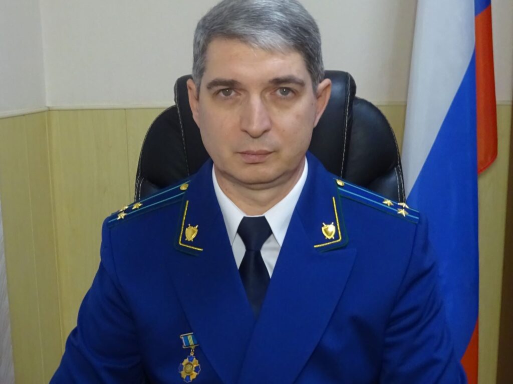 Мужчина в синем форменном кителе с погонами и наградой сидит в кресле на фоне российского флага