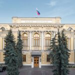 Фасад здания Банка России с российским флагом на крыше и хвойными деревьями на переднем плане
