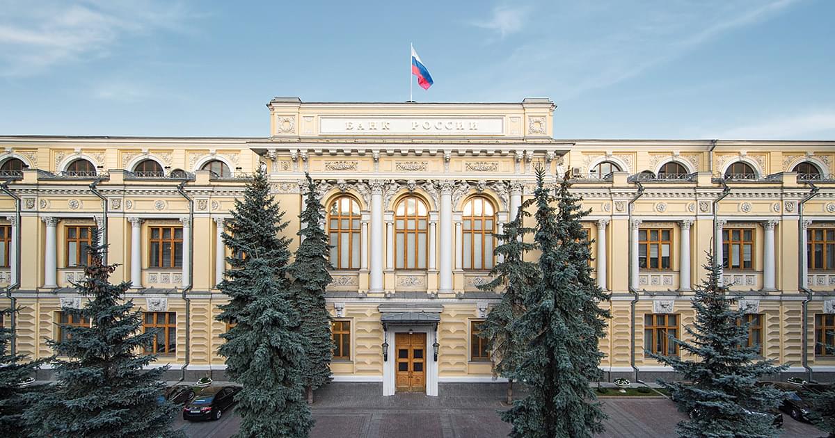 Фасад здания Банка России с российским флагом на крыше и хвойными деревьями на переднем плане