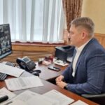 мужчина в офисе за столом смотрит на экран с сеткой участников видеоконференции; на столе документы, телефон и оргтехника
