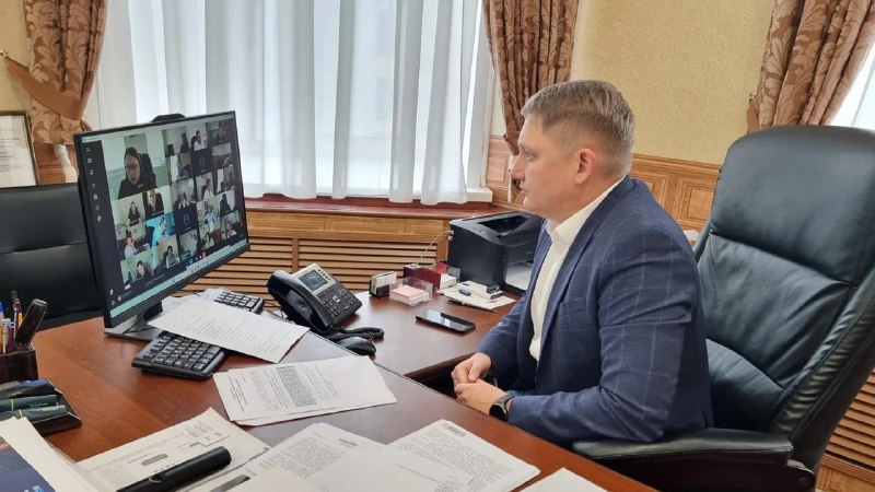 мужчина в офисе за столом смотрит на экран с сеткой участников видеоконференции; на столе документы, телефон и оргтехника