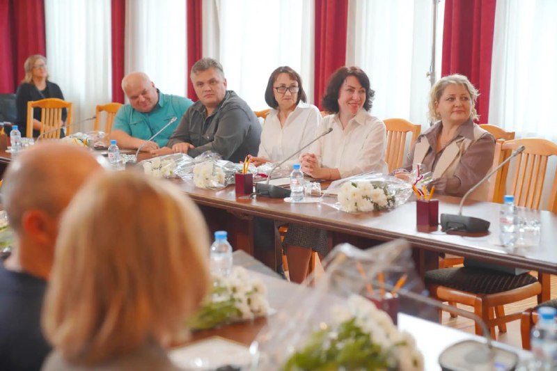 Несколько людей за длинным столом в зале заседаний; на столах букеты, бутылки с водой и микрофоны.