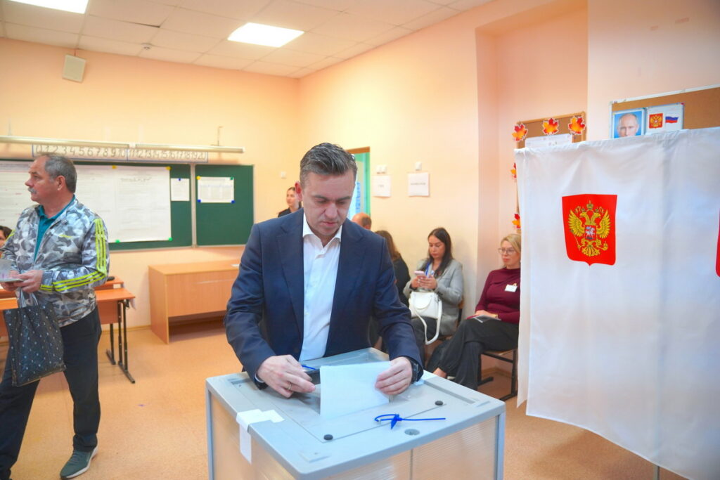 Мужчина в костюме опускает бюллетень в прозрачную урну рядом с кабиной для голосования