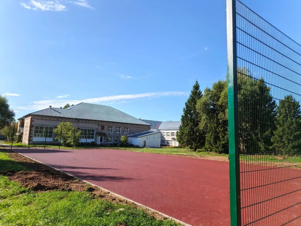 Спортивная площадка с красным покрытием и высоким сетчатым ограждением рядом с кирпичным зданием