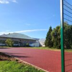 Спортивная площадка с красным покрытием и высоким сетчатым ограждением рядом с кирпичным зданием