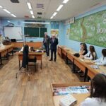Учебный класс: школьники сидят за партами, два пожилых мужчины (ветераны) стоят у переднего стола с предметами из экспозиции (фуражка, каска, атрибуты), на столах — листовки о полиции.