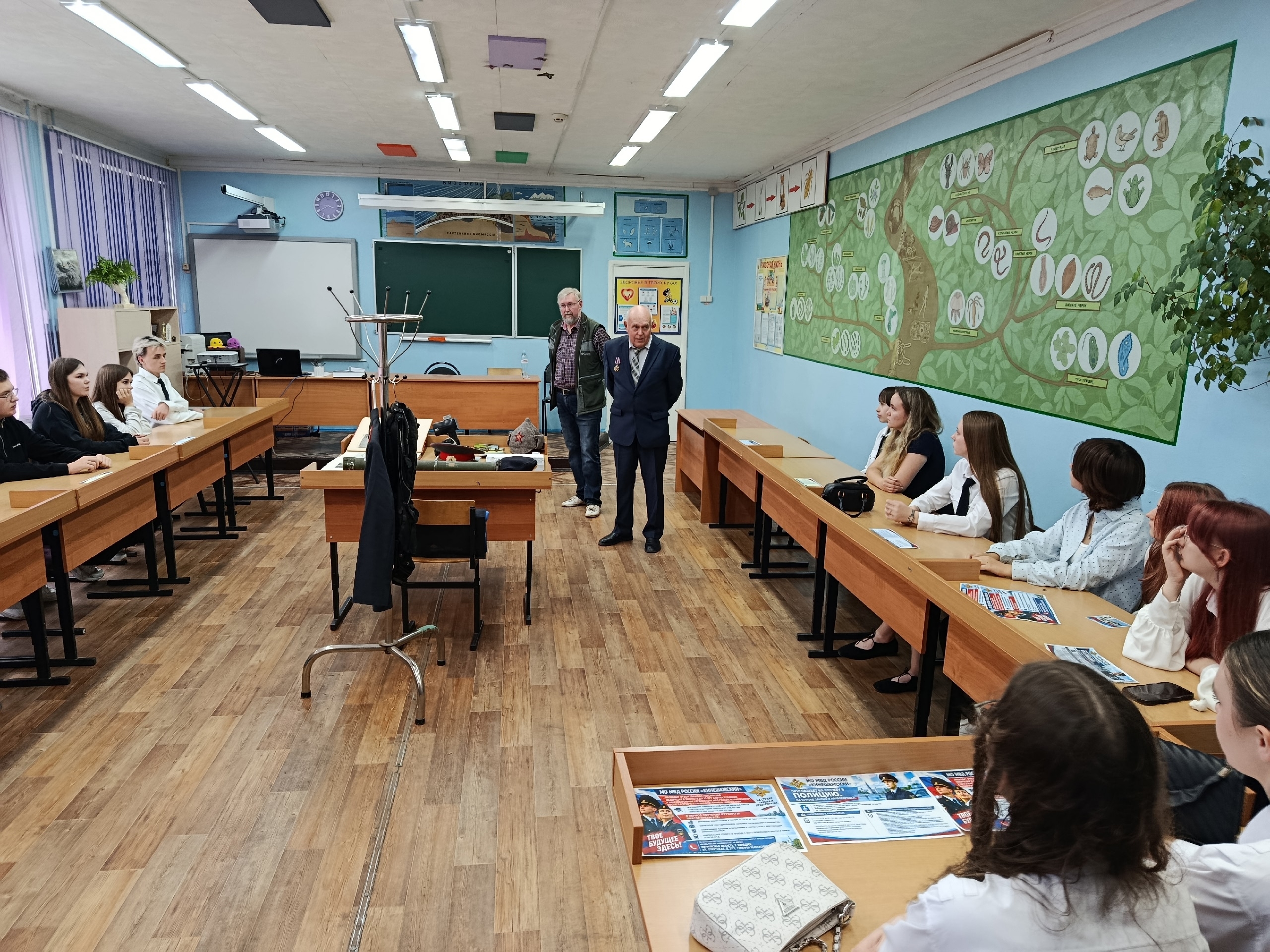 Учебный класс: школьники сидят за партами, два пожилых мужчины (ветераны) стоят у переднего стола с предметами из экспозиции (фуражка, каска, атрибуты), на столах — листовки о полиции.