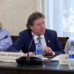 Мужчина в костюме за столом на совещании; перед ним микрофон, бутылки с водой и настольная табличка с фамилией.
