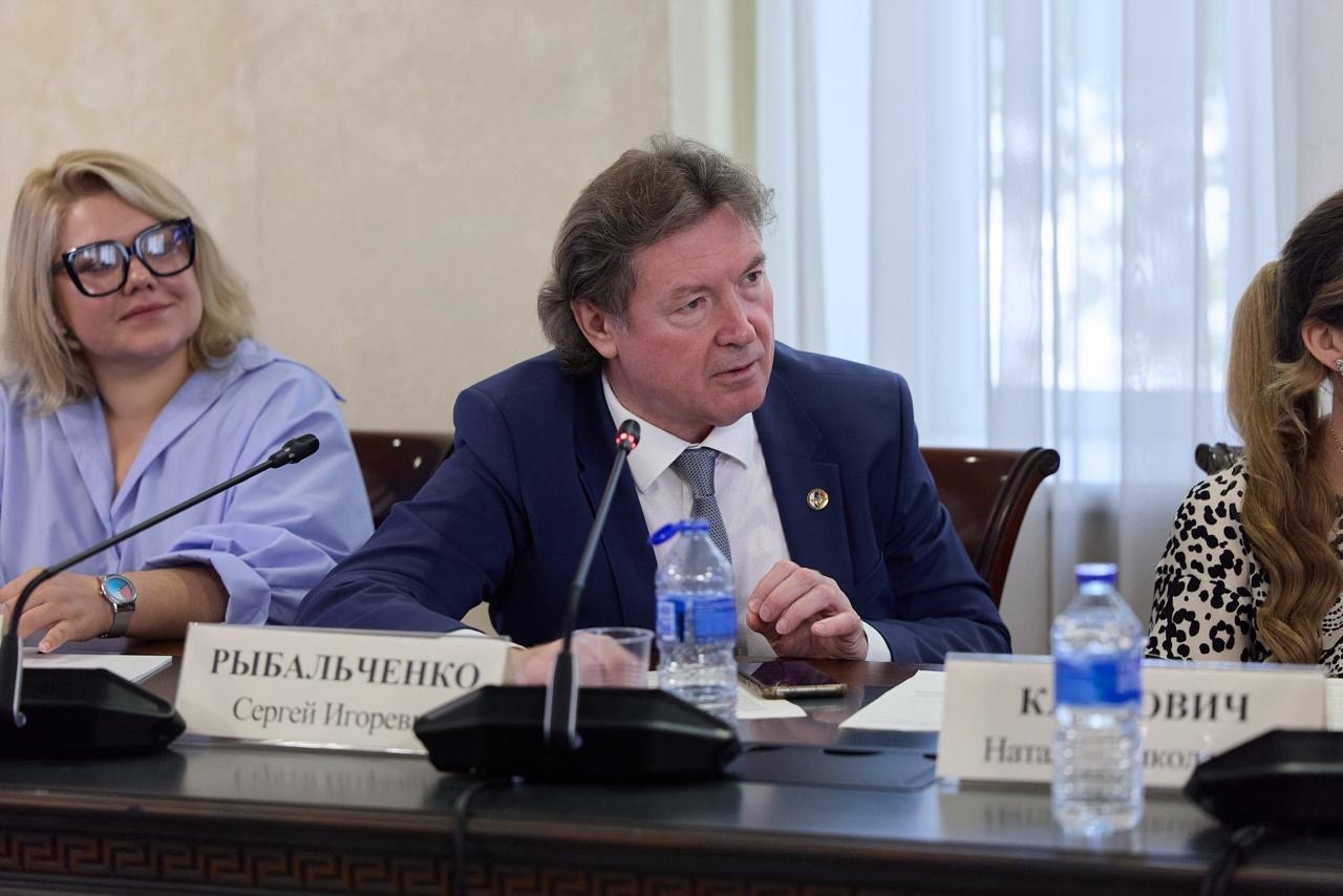 Мужчина в костюме за столом на совещании; перед ним микрофон, бутылки с водой и настольная табличка с фамилией.