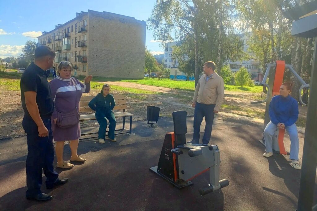 Группа взрослых беседует у тренажёров и скамейки на спортивной площадке среди жилых домов