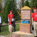 мемориальный знак на кирпичной кладке с табличкой и механическими весами на верхней полке; рядом дети и взрослые в ходе церемонии