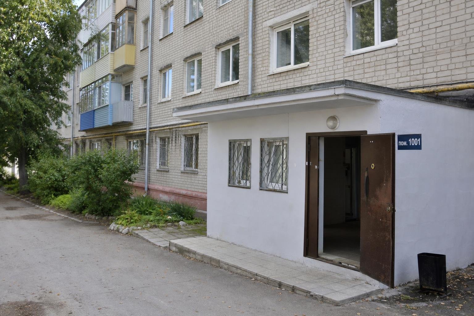 Фасад пятиэтажного кирпичного жилого дома с пристроенным входом в нежилое помещение; открытая дверь и синяя табличка с надписью «пом. 1001».