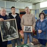 Четверо человек в музейном интерьере держат в руках две рамки с чёрно‑белыми фотографиями (одна большая портретная), на стенах — музейные стенды и экспонаты.