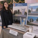 студентка у выставочного стола с макетом и постерами проекта въездного знака «Наволоки»