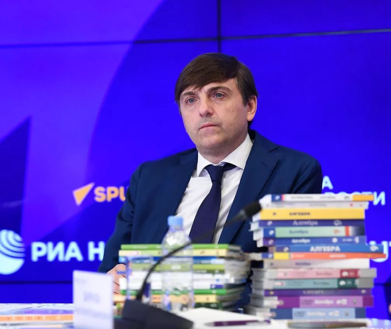 мужчина в тёмном костюме сидит за столом на фоне баннера «РИА Новости», перед ним несколько стопок учебников и микрофон