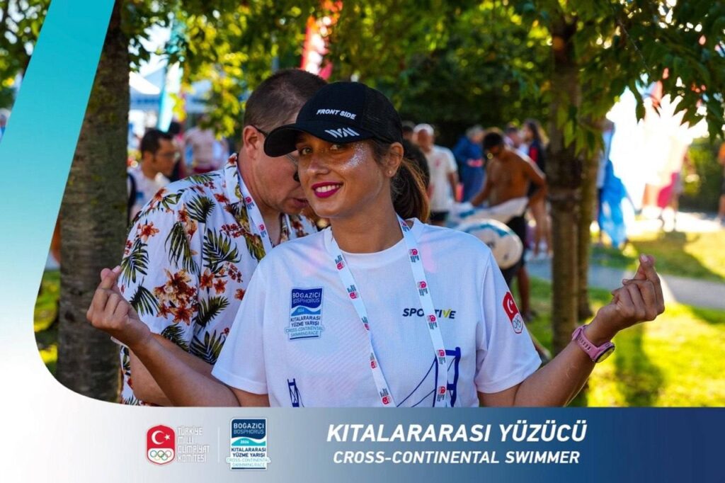 Улыбающаяся женщина в спортивной футболке и чёрной кепке позирует за рамкой с надписью «Cross-continental swimmer».
