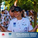 Улыбающаяся женщина в спортивной футболке и чёрной кепке позирует за рамкой с надписью «Cross-continental swimmer».