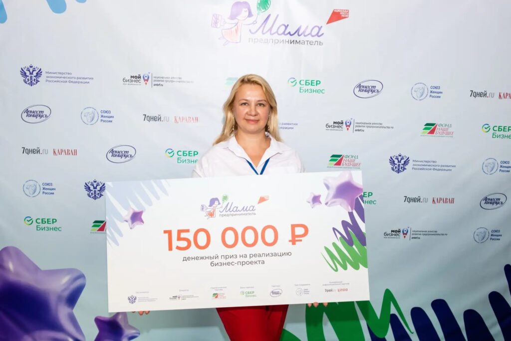 Женщина держит большой сертификат на денежный приз в 150 тысяч рублей на фоне стены с логотипами