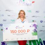 Женщина держит большой сертификат на денежный приз в 150 тысяч рублей на фоне стены с логотипами