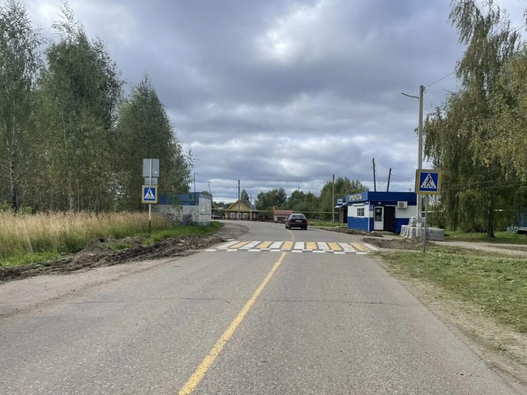 Дорога с пешеходным переходом, автомобилем и разрытыми обочинами; справа небольшое здание и стройматериалы