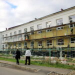 Рабочие утепляют фасад двухэтажного здания со строительных лесов, двое прохожих идут вдоль ограждения