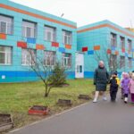 яркий фасад двухэтажного детского сада с цветной облицовкой; на дорожке группа детей в зимней одежде под присмотром взрослого