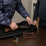 Росгвардеец рассматривает ружьё в чехле на столе, рядом патроны; видны руки двух мужчин