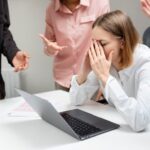 Подавленная сотрудница за столом с ноутбуком; рядом коллеги жестами выражают недовольство.